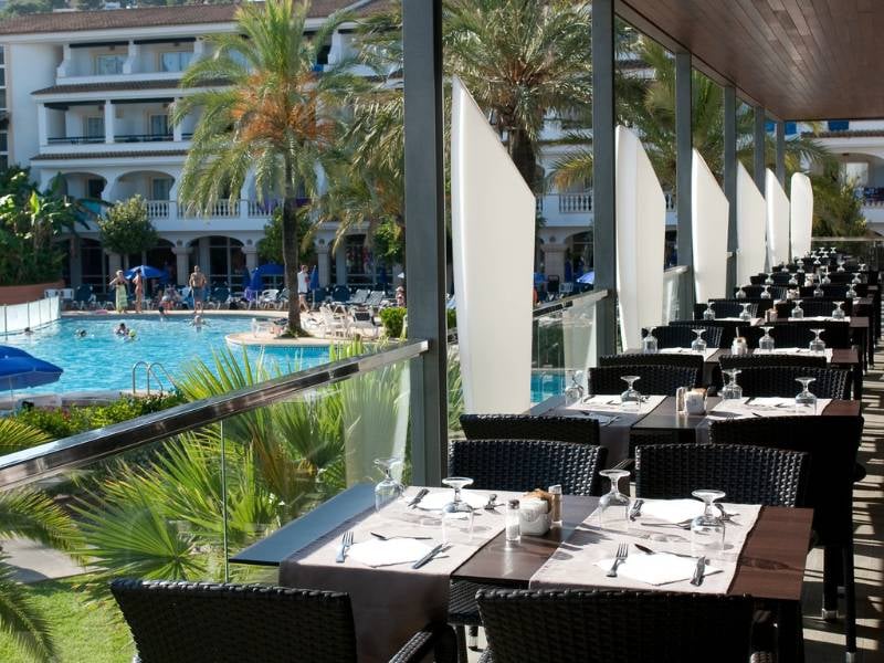 Outdoor dining area at a beach club restaurant with tables overlooking a pool and palm trees.