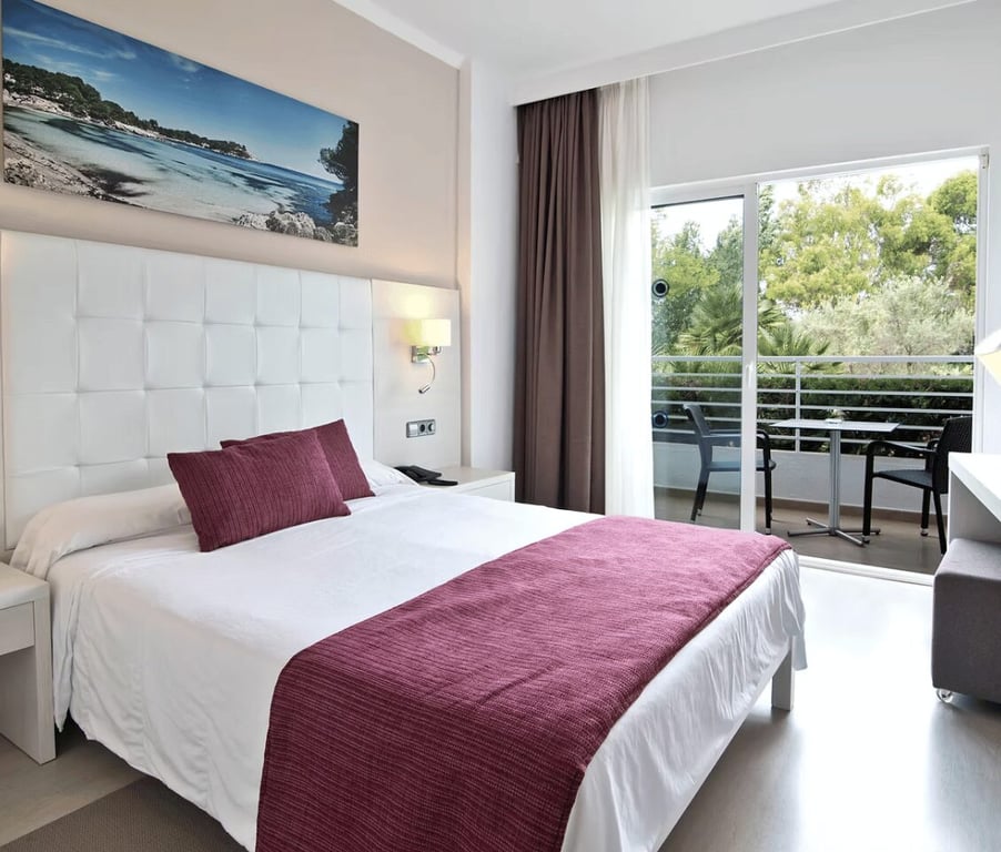 Modern beach club room with a double bed, burgundy bedding, white headboard, landscape artwork, and a balcony with lush green views.