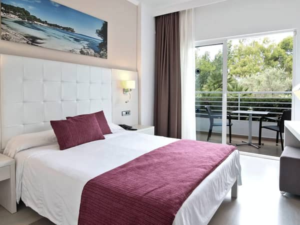 Modern beach club room with a double bed, burgundy bedding, white headboard, landscape artwork, and a balcony with lush green views.