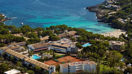 Aerial view of a beach club with tennis courts, swimming pools, and hotel buildings next to a beautiful bay and lush greenery.