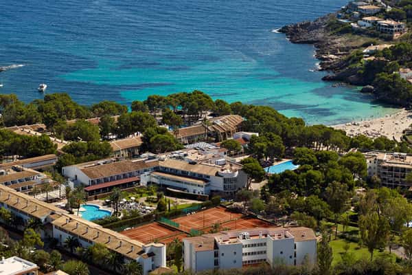 Aerial view of a beach club with tennis courts, swimming pools, and hotel buildings next to a beautiful bay and lush greenery.