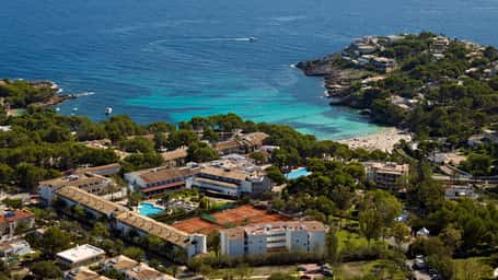 Aerial view of a beach club with resort facilities, tennis courts, pool, and coastline with turquoise waters and sandy beaches.