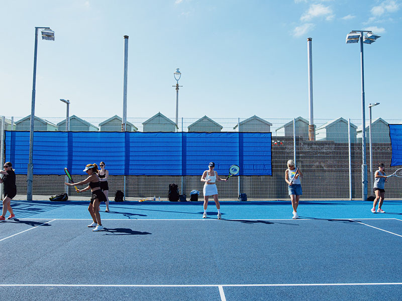 Outdoor tennis match in Brighton with players on a blue court, seaside beach huts in the background under a clear sky.