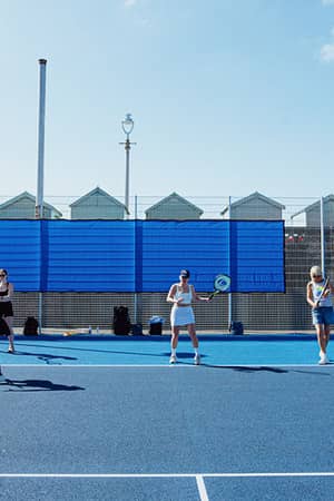 Outdoor tennis match in Brighton with players on a blue court, seaside beach huts in the background under a clear sky.