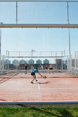 People playing padel tennis on a fenced outdoor court in Brighton with blue skies overhead and modern houses in the background.