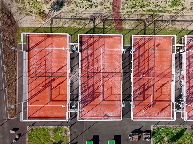 Aerial view of three orange padel courts with players, surrounded by greenery, during a tennis padel weekend in Brighton.