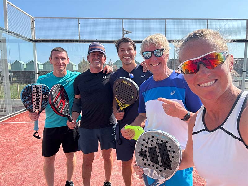 Five people standing on a red padel court, each holding a padel racket, on a sunny day in Brighton.