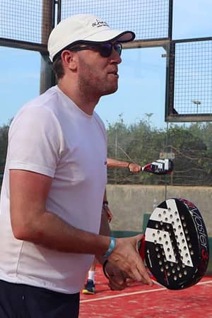 Man wearing a white t-shirt and cap, holding a padel racquet on a court in Brighton, ready to play.