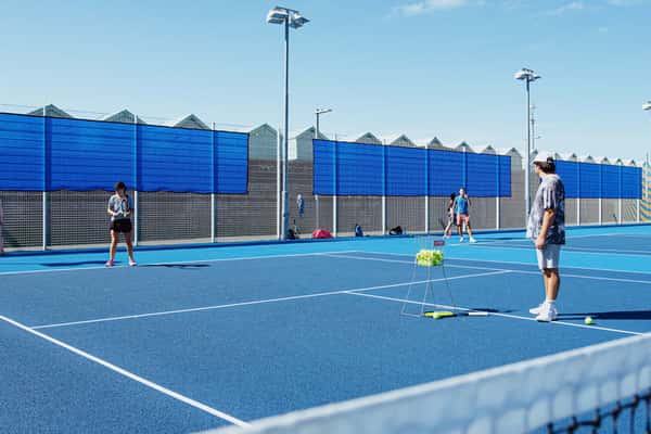 Players on a blue padel tennis court in Brighton, surrounded by fencing and light towers under a clear sky, with a basket of tennis balls at hand.