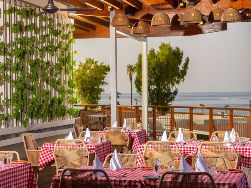 Outdoor seating at a beach restaurant with red and white checkered tablecloths, sea view, and greenery on the wall.