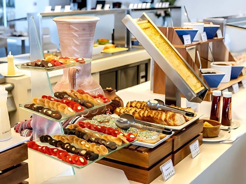 Buffet arrangement at Capo Bay Gallery showcasing assorted pastries, sandwiches, and breads on glass stands with condiments in the background.