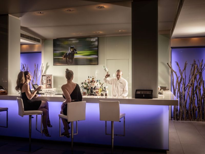Two women in elegant attire sit at Capo Bay's evening bar, with a bartender preparing a drink and a nature scene on the screen.