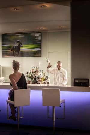 Two women in elegant attire sit at Capo Bay's evening bar, with a bartender preparing a drink and a nature scene on the screen.