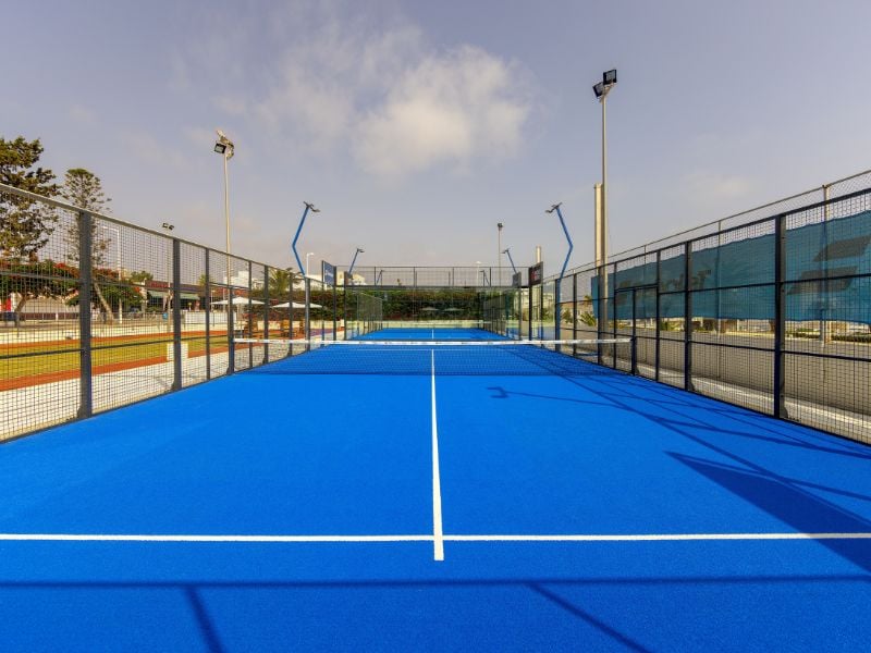 Blue padel court with wire fencing and floodlights at Capo Bay Gallery, set under a clear sky.