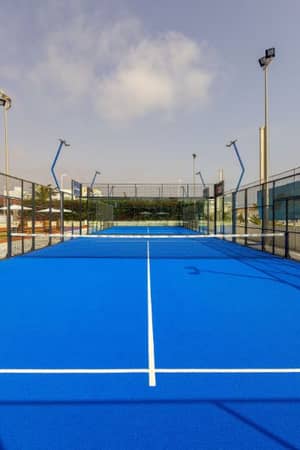 Blue padel court with wire fencing and floodlights at Capo Bay Gallery, set under a clear sky.