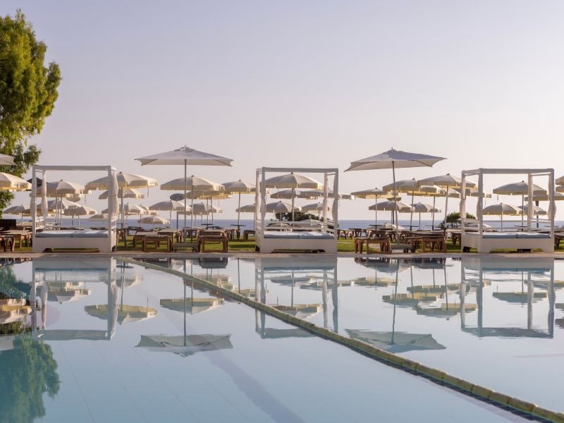 Capo Bay hotel pool exterior with canopied sunbeds and parasols reflected in the water, against a clear sky in Protaras, Cyprus.