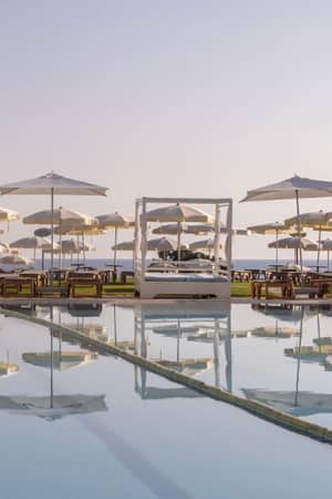 Capo Bay hotel pool exterior with canopied sunbeds and parasols reflected in the water, against a clear sky in Protaras, Cyprus.