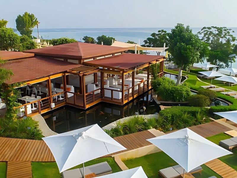 Aerial view of Capo Bay Gallery restaurant with wooden architecture, surrounded by greenery and sea, featuring outdoor seating under white umbrellas.