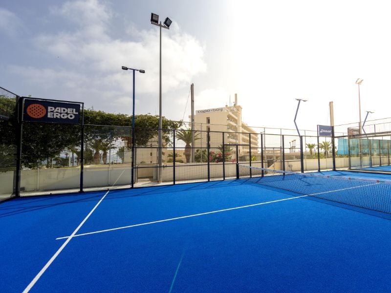 Outdoor padel courts with blue flooring, surrounded by metal fencing, under a sunny sky at Capo Bay Gallery.