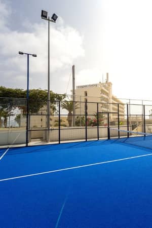 Outdoor padel courts with blue flooring, surrounded by metal fencing, under a sunny sky at Capo Bay Gallery.