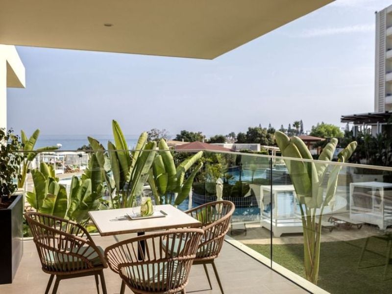 A veranda at Capo Bay Hotel with wicker chairs and a table, overlooking tropical plants and the sea in the background.