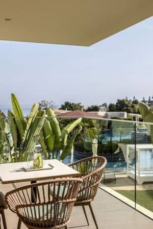 A veranda at Capo Bay Hotel with wicker chairs and a table, overlooking tropical plants and the sea in the background.