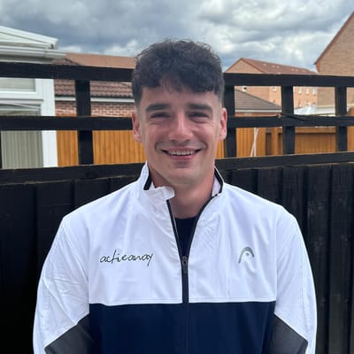 Cameron smiling outdoors, wearing an 'achievancy' branded blue and white jacket, with a fence and cloudy sky in the background.