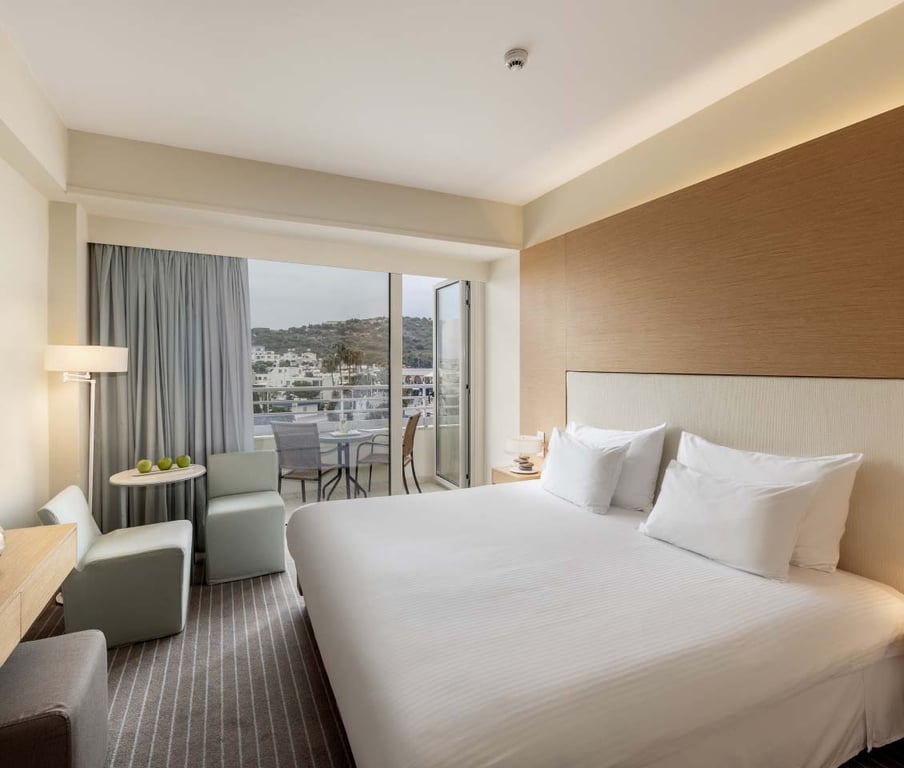 Hotel room with a double bed, neutral decor, and a balcony offering an inland view of hills and buildings.