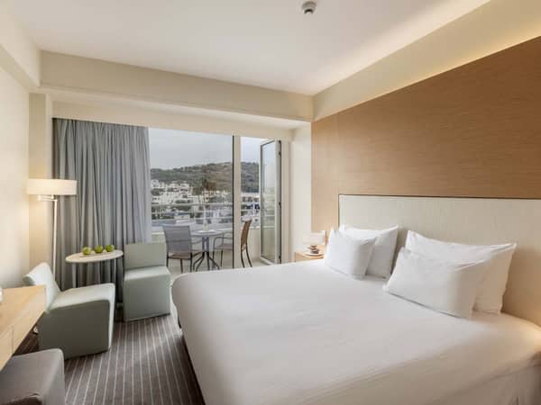 Hotel room with a double bed, neutral decor, and a balcony offering an inland view of hills and buildings.