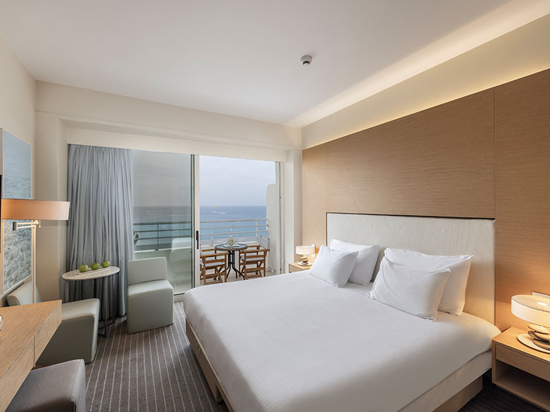 Luxurious Capo Bay hotel room with modern design and a sea view balcony, featuring a large bed, soft lighting, and a dining area.