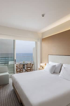 Luxurious Capo Bay hotel room with modern design and a sea view balcony, featuring a large bed, soft lighting, and a dining area.