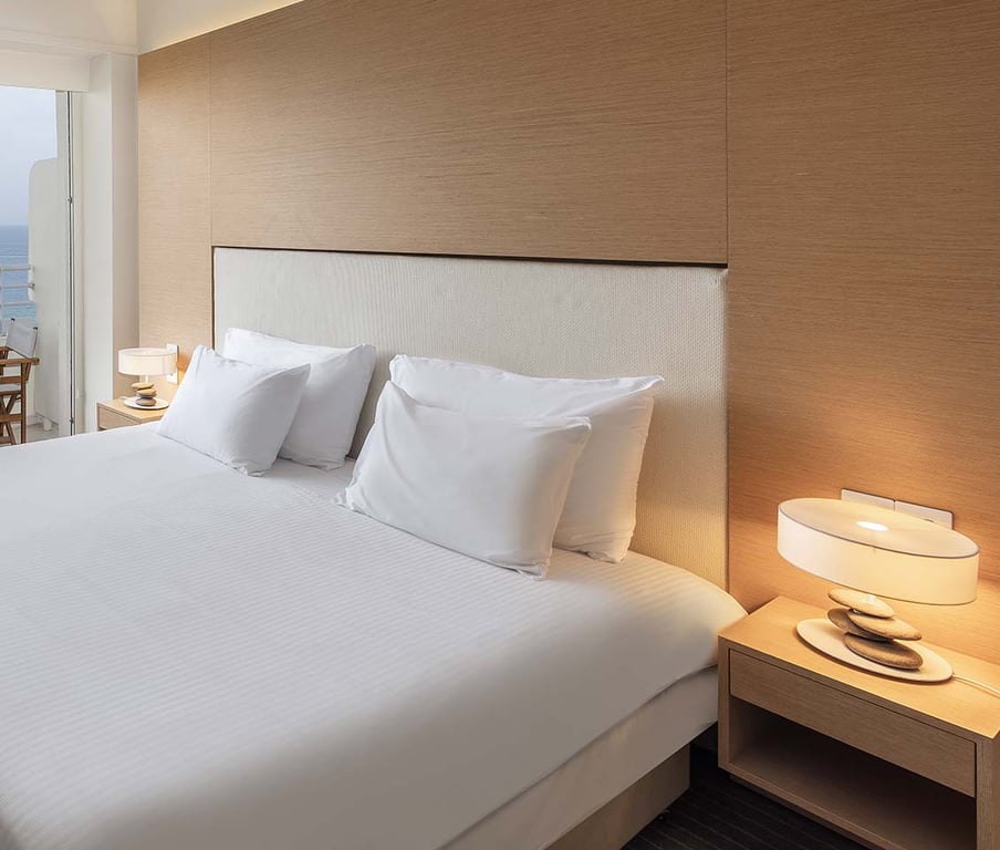 Modern hotel room with a large bed, crisp white linens, bedside tables with lamps, and a side sea view balcony.