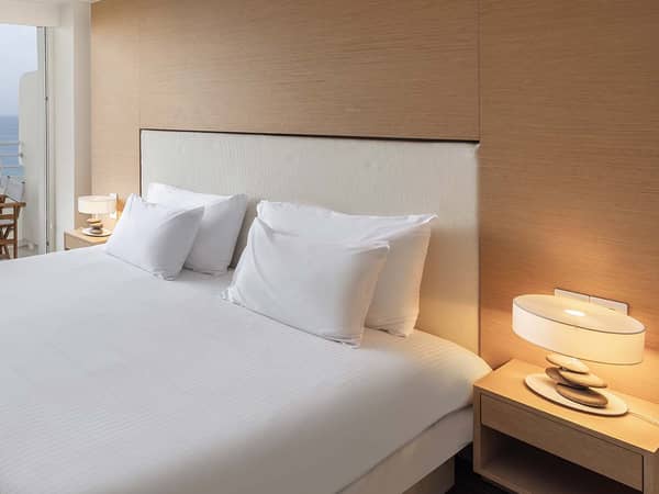 Modern hotel room with a large bed, crisp white linens, bedside tables with lamps, and a side sea view balcony.