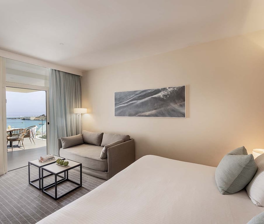 Hotel room with modern decor, comfortable bed, grey sofa, and a balcony with sea views.