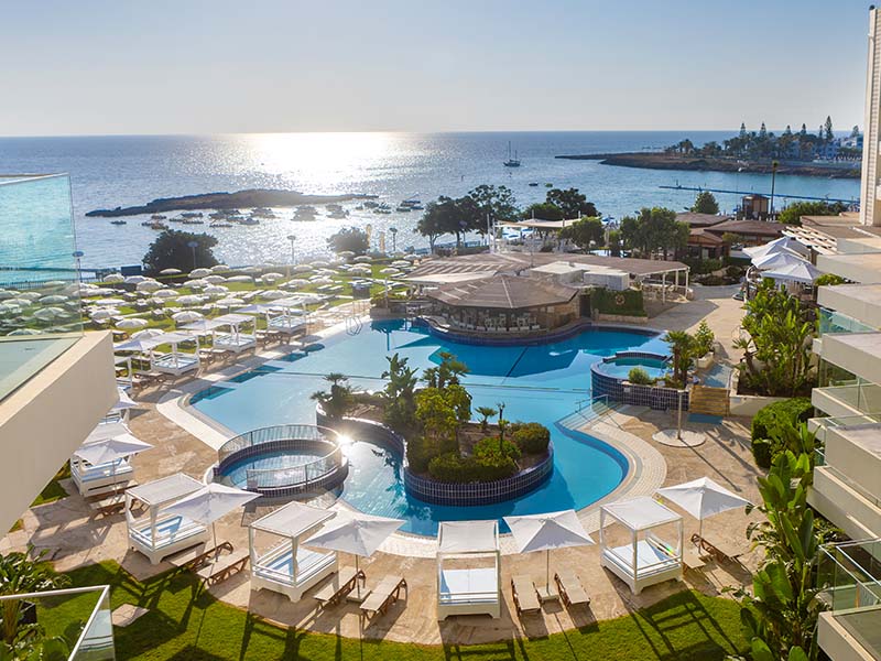 Overhead view of Capo Bay Hotel's pool area with sun loungers, umbrellas, and the Mediterranean Sea in Protaras, Cyprus in the background.