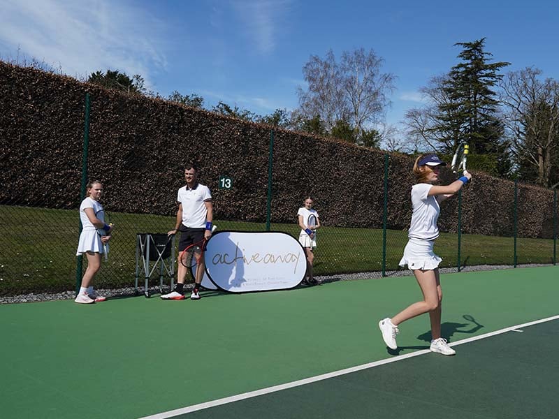 Jamie Murray on a tennis court with junior students at Churcher's School, during a training session with an ACE ACADEMY banner in the background.