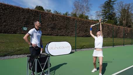 Jamie Murray coaching a student on a grass tennis court at Churcher's College, with the student poised to hit the ball.