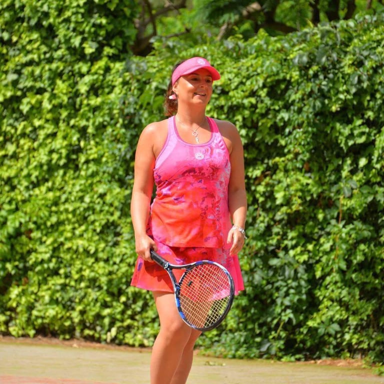 Claudia Agha wearing a pink tennis outfit, holding a tennis racket on a sunlit court with greenery in the background.