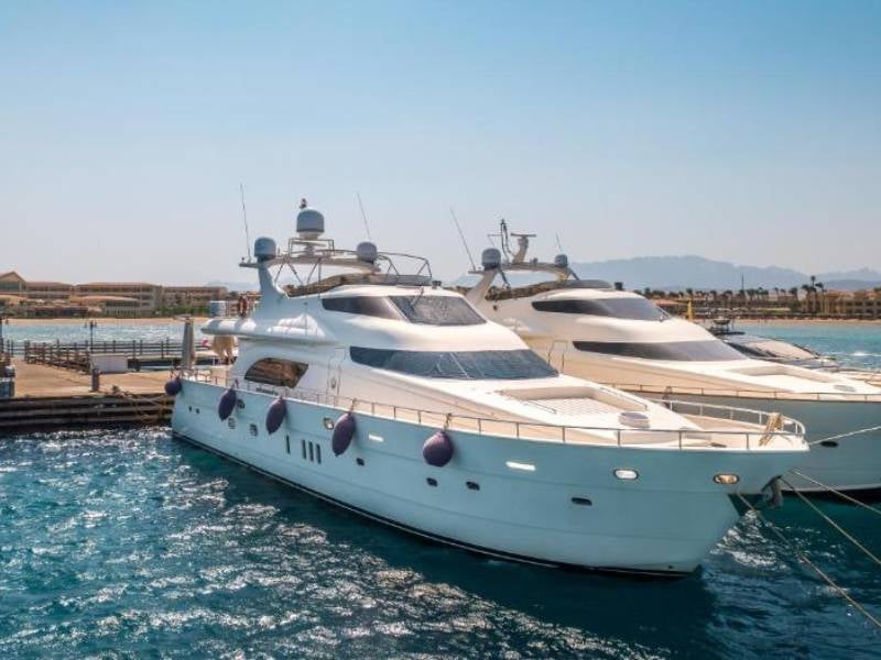 Two white motor yachts docked at a marina, showcasing sleek designs and luxurious features, with a clear sky and distant mountains in the background.