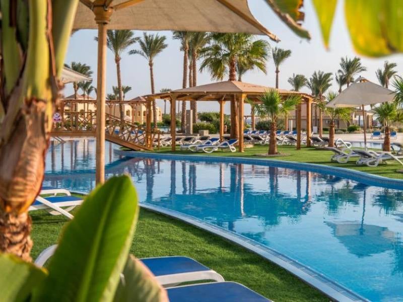 Cleopatra Luxury Resort pool area with blue pool, sun loungers, umbrellas, green lawns, and palm trees in a tranquil setting.