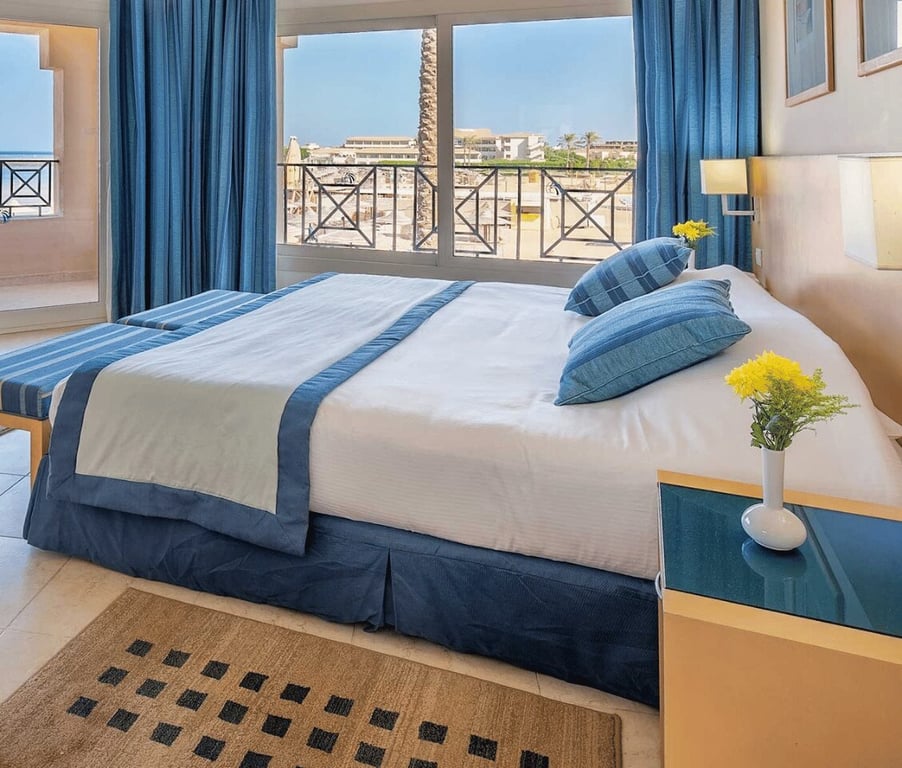 Luxurious Cleopatra Junior Suite featuring a king-sized bed with blue and white bedding, large windows, and a vase with yellow flowers on a side table.