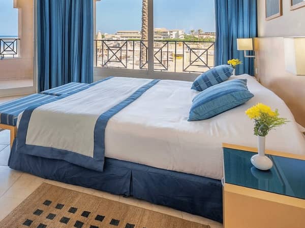 Luxurious Cleopatra Junior Suite featuring a king-sized bed with blue and white bedding, large windows, and a vase with yellow flowers on a side table.
