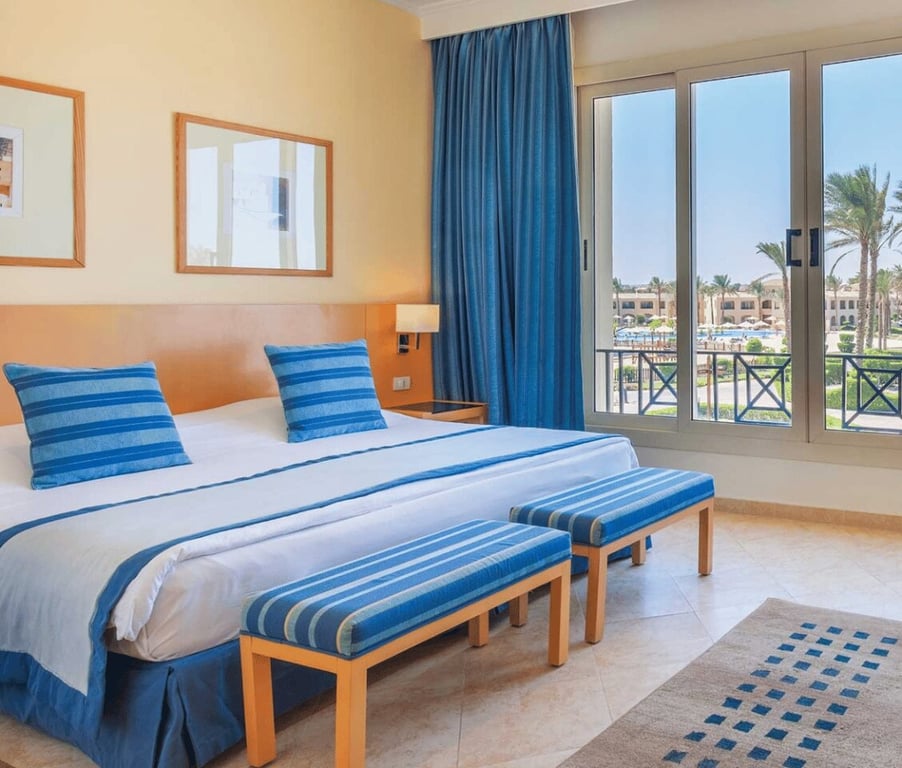 Interior of the Cleopatra Master Suite with a king-sized bed, blue-striped decor, and large windows overlooking palm trees and a sunny sky.