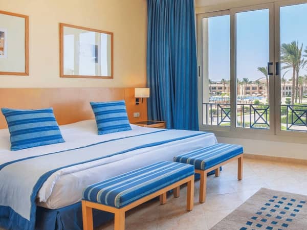 Interior of the Cleopatra Master Suite with a king-sized bed, blue-striped decor, and large windows overlooking palm trees and a sunny sky.