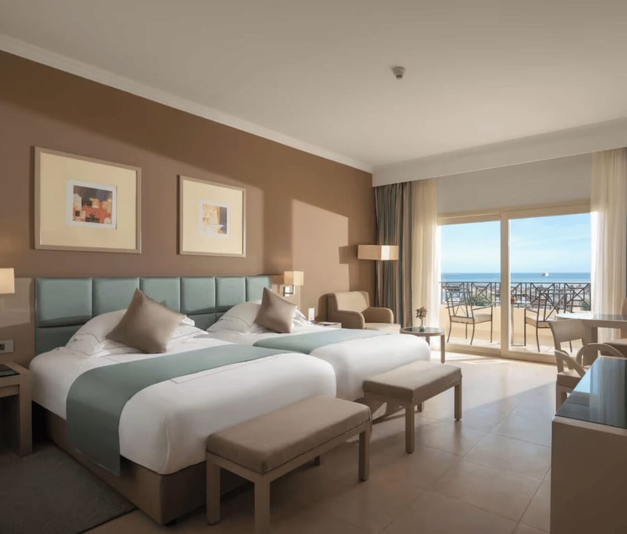 Luxurious hotel room with two double beds, taupe and teal decor, balcony with sea view, modern furnishings, and contemporary art.