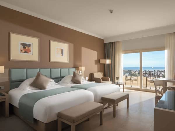 Luxurious hotel room with two double beds, taupe and teal decor, balcony with sea view, modern furnishings, and contemporary art.