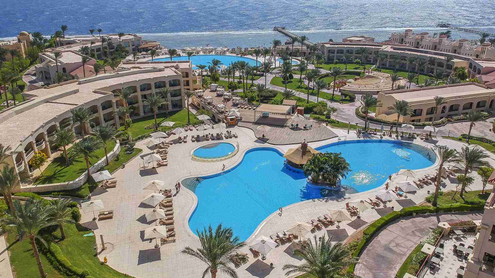 Aerial view of Cleopatra Resort, highlighting a large central swimming pool, poolside loungers, and surrounding resort buildings with a stunning ocean backdrop.