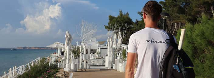 Man in 'achievamind' T-shirt walking towards a seaside resort with a tennis bag and racquet, surrounded by trees and overlooking the ocean.