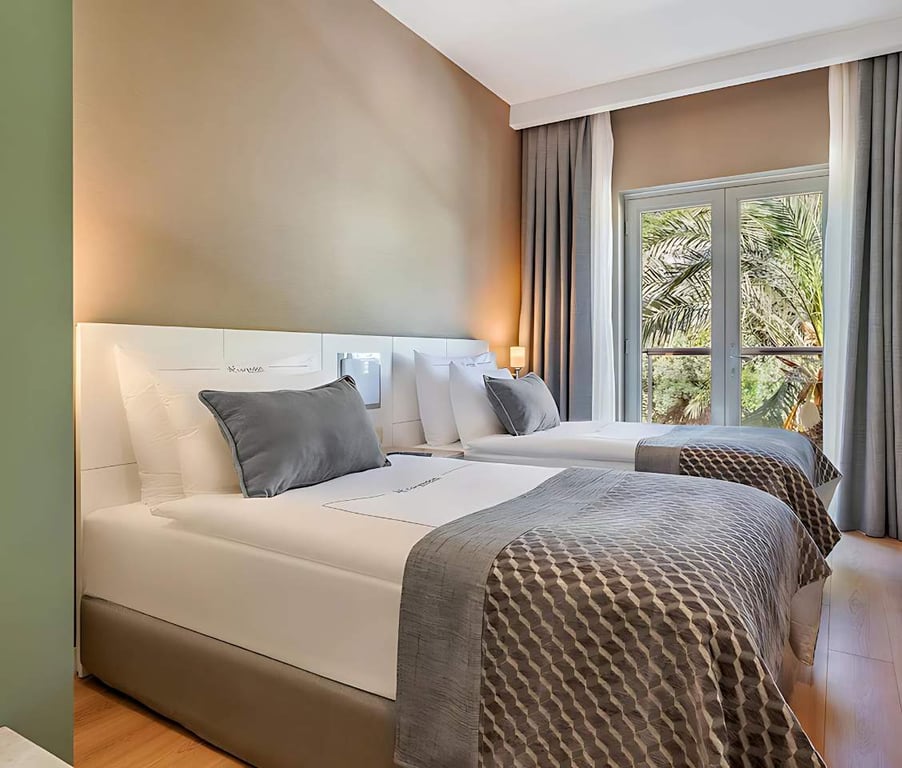 Hotel family room with two double beds, grey and white bedding, and a window showing palm trees outside.