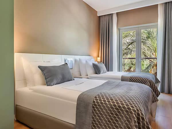 Hotel family room with two double beds, grey and white bedding, and a window showing palm trees outside.